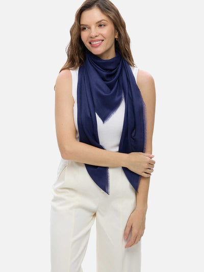A woman modeling a navy blue Featherlight Square Cashmere Scarf wrapped around her neck over a white sleeveless top and cream-colored pants, set against a solid light gray background.
