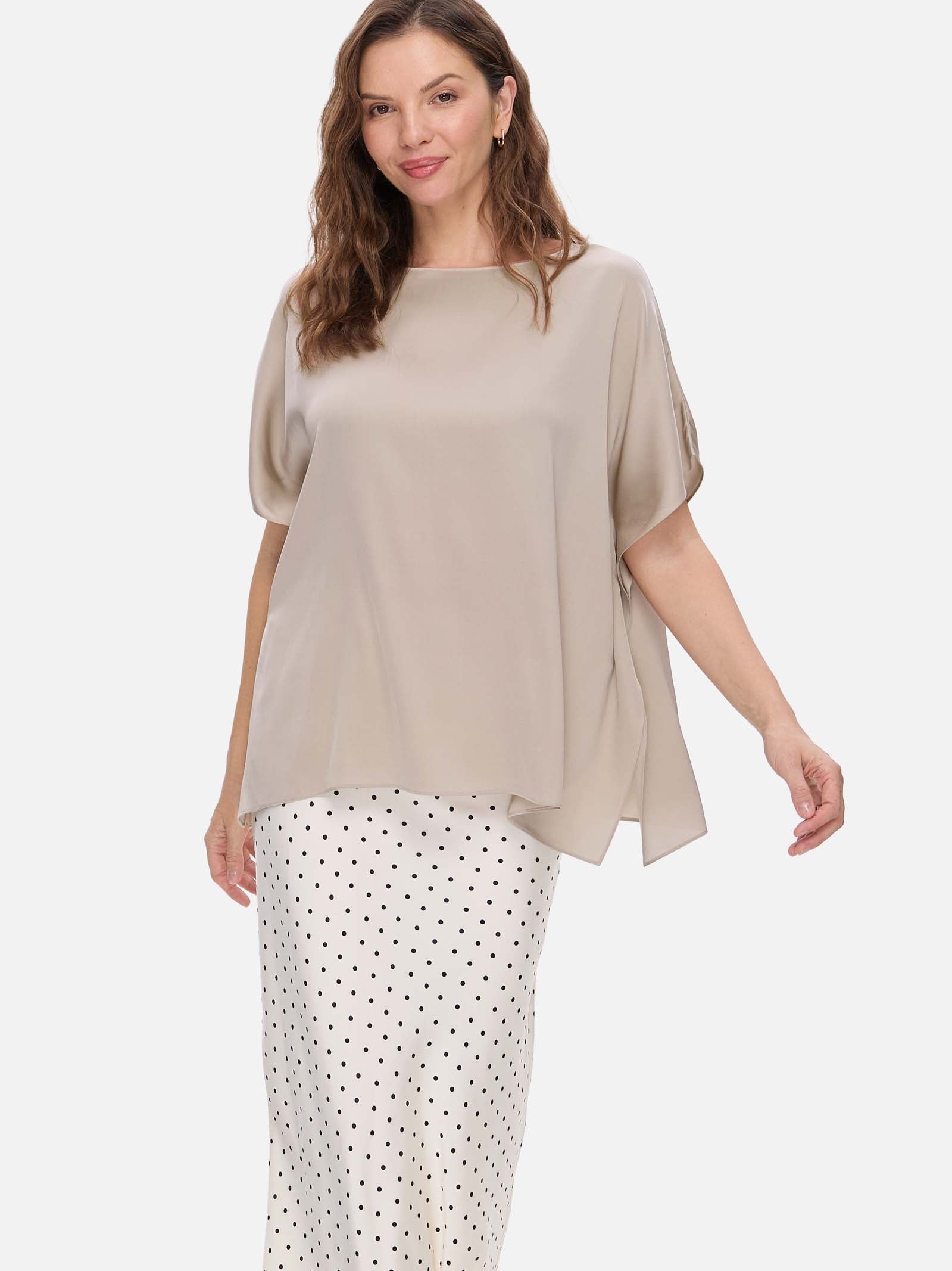 A woman modeling a loose, short-sleeve taupe silk top paired with white pants featuring a black polka-dot pattern, set against a plain light gray studio background.