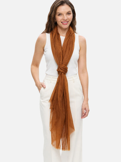 A woman modeling a featherlight chocolate cashmere scarf tied around her neck, paired with a white sleeveless top and white pants, against a light gray studio background.