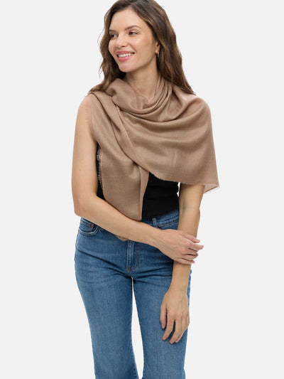 A woman modeling a camel Featherlight Square Cashmere Scarf draped over her shoulders, styled with a black top and blue jeans against a plain light gray studio background.