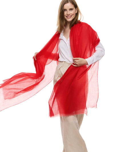 A woman modeling a red featherlight cashmere scarf draped over her shoulders, paired with a white blouse and beige trousers, set against a solid white background.