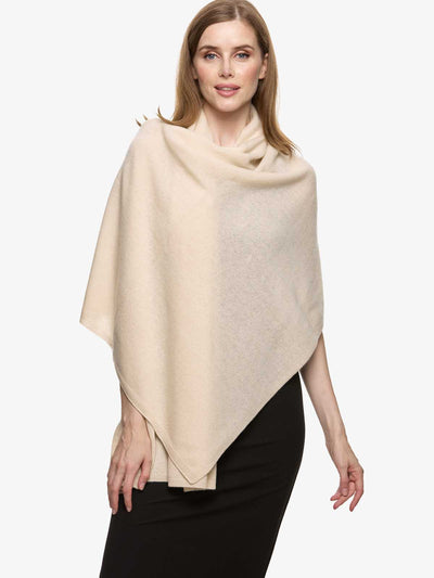 A woman modeling a large, soft triangular cream-colored cashmere wrap or poncho draped over her shoulders, paired with a black skirt, against a plain light gray studio background.