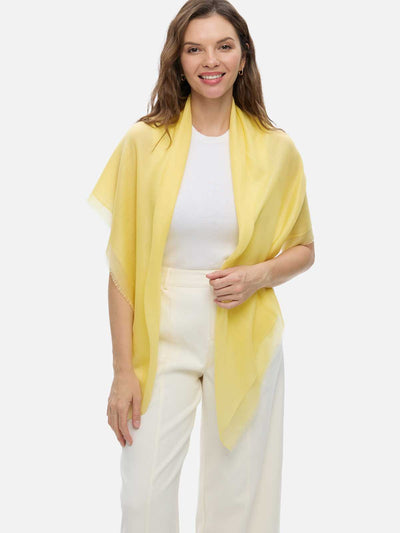 A woman modeling a Featherlight Square Yellow Cashmere Scarf draped over her shoulders, styled over a white top and cream-colored trousers, against a solid light gray background.