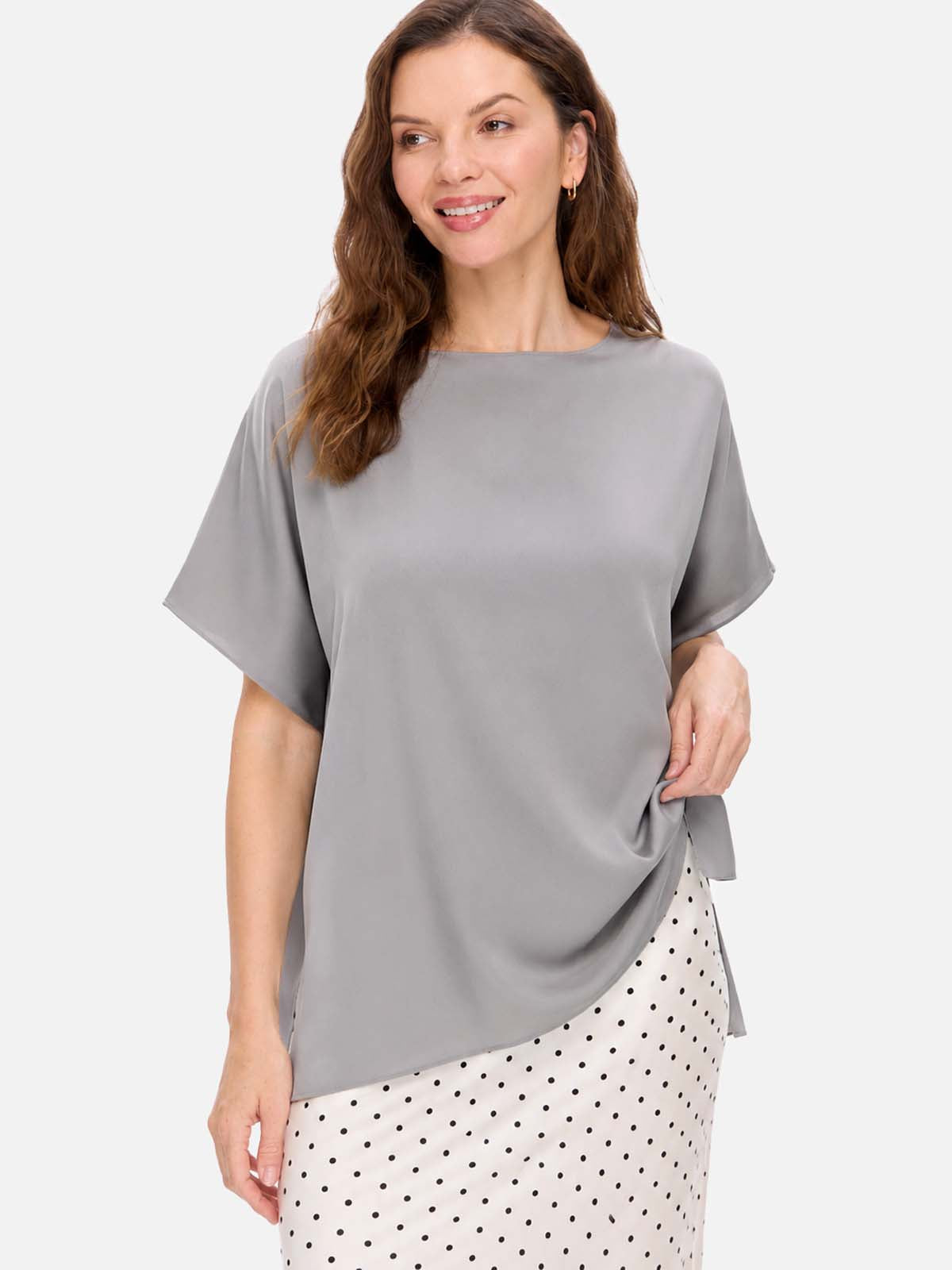 A woman wearing a loose, short-sleeve gray silk top and a white skirt with black polka dots, photographed against a light gray studio background.