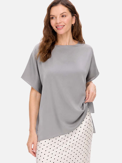 A woman wearing a loose, short-sleeve gray silk top and a white skirt with black polka dots, photographed against a light gray studio background.