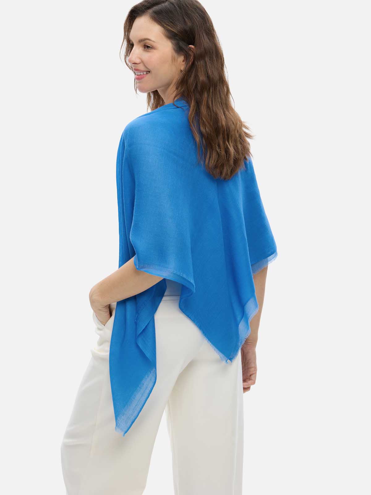 A woman wearing a blue Featherlight Square Cashmere Scarf draped over a white sleeveless top and cream-colored trousers, photographed against a light gray studio background.