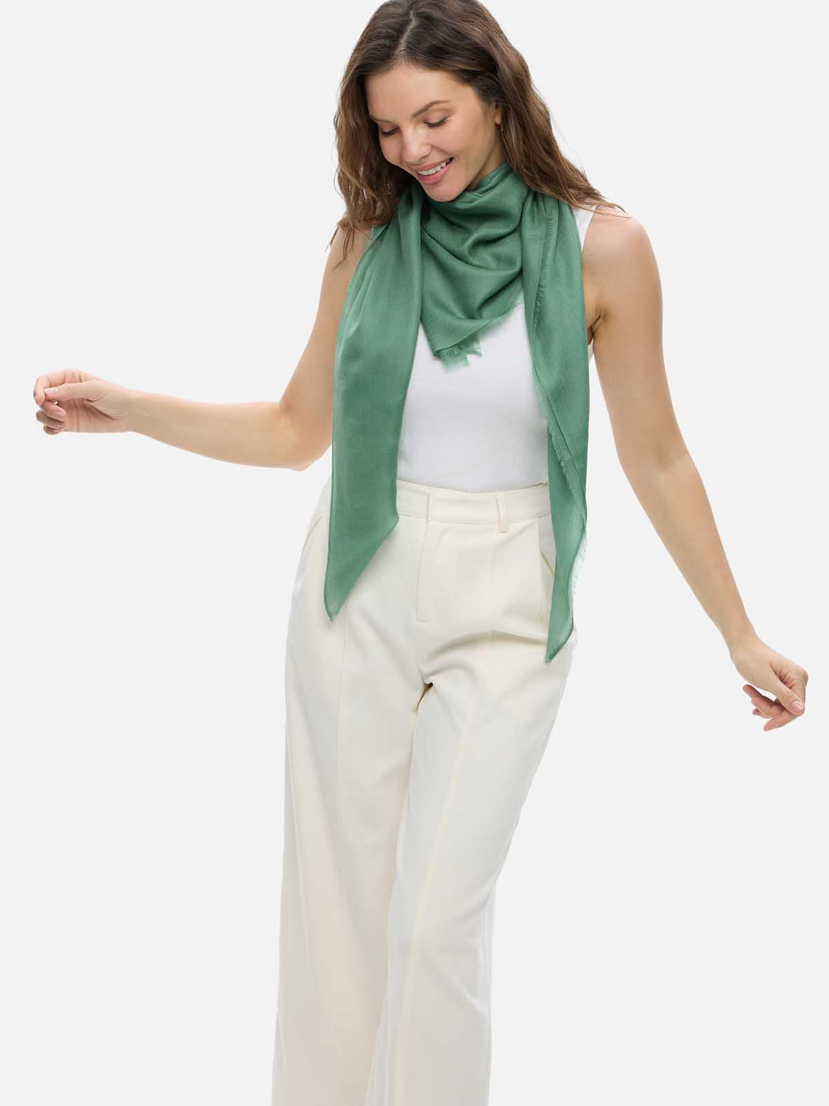 A woman wearing a sage green cashmere scarf draped over her shoulders, paired with a white top and cream-colored trousers, against a solid light gray background.