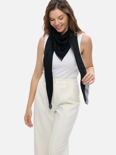 A woman modeling a lightweight black cashmere scarf wrapped around her neck over a white sleeveless top and cream-colored trousers, set against a solid light gray background.