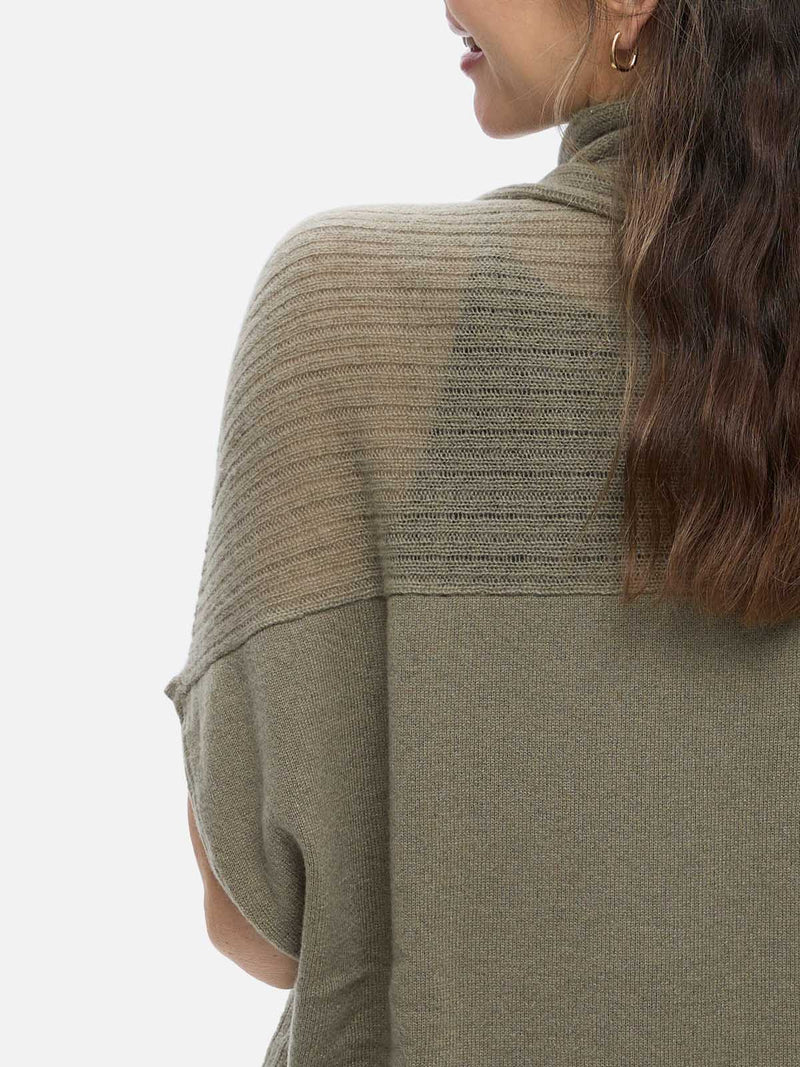 Woman wearing a green cashmere vest over a black top and blue jeans on a white background