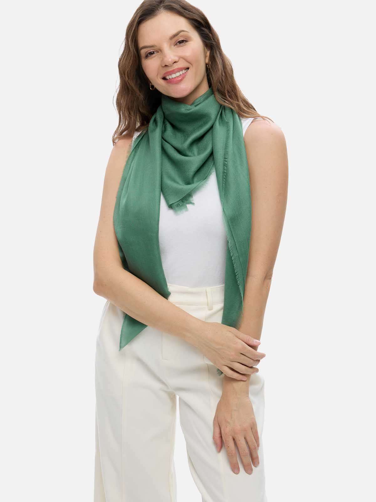 A woman wearing a sage green cashmere scarf, paired with a white top and cream-colored trousers, against a solid light gray background.