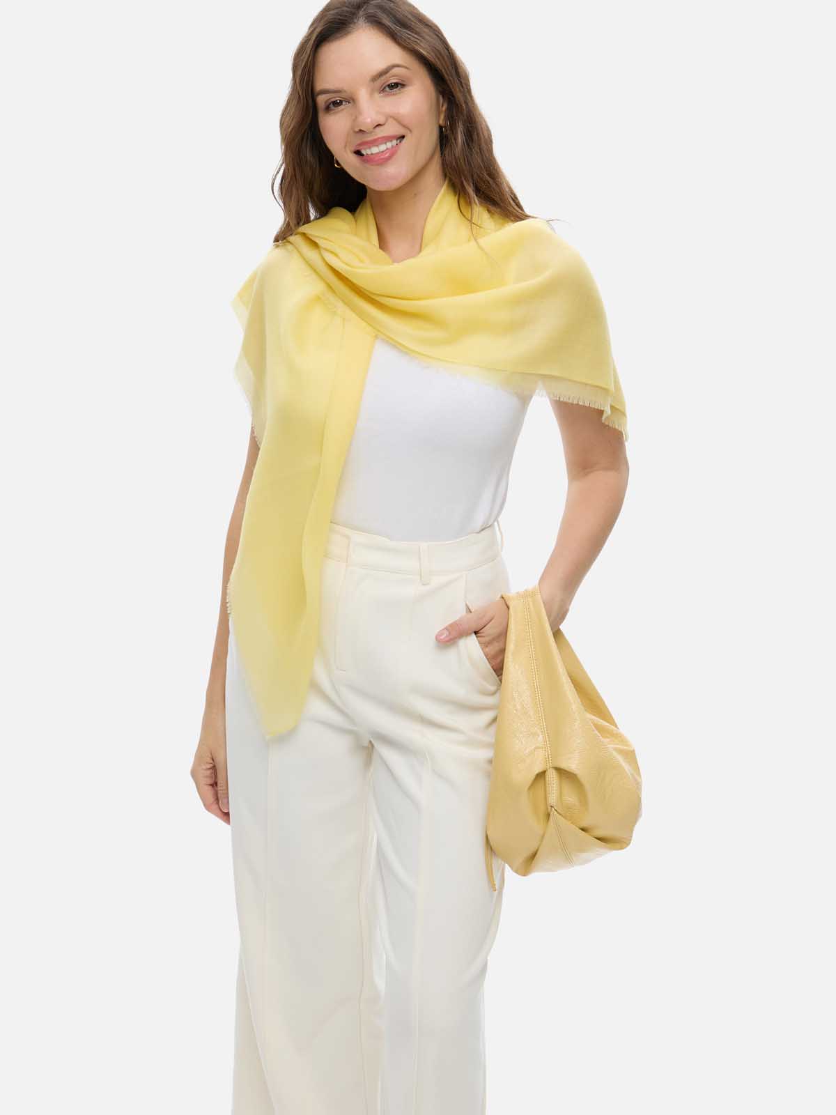 A woman modeling a Featherlight Square Yellow Cashmere Scarf draped over her shoulders, styled over a white top and cream-colored trousers, against a solid light gray background.