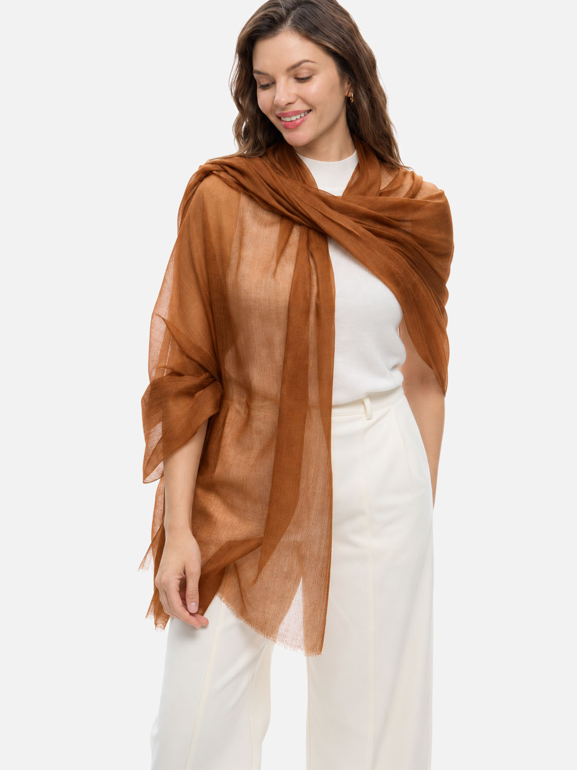 A woman wearing a lightweight, semi-sheer featherlight chocolate cashmere scarf draped over her shoulders, styled over a white top and white trousers, set against a plain light gray studio background.