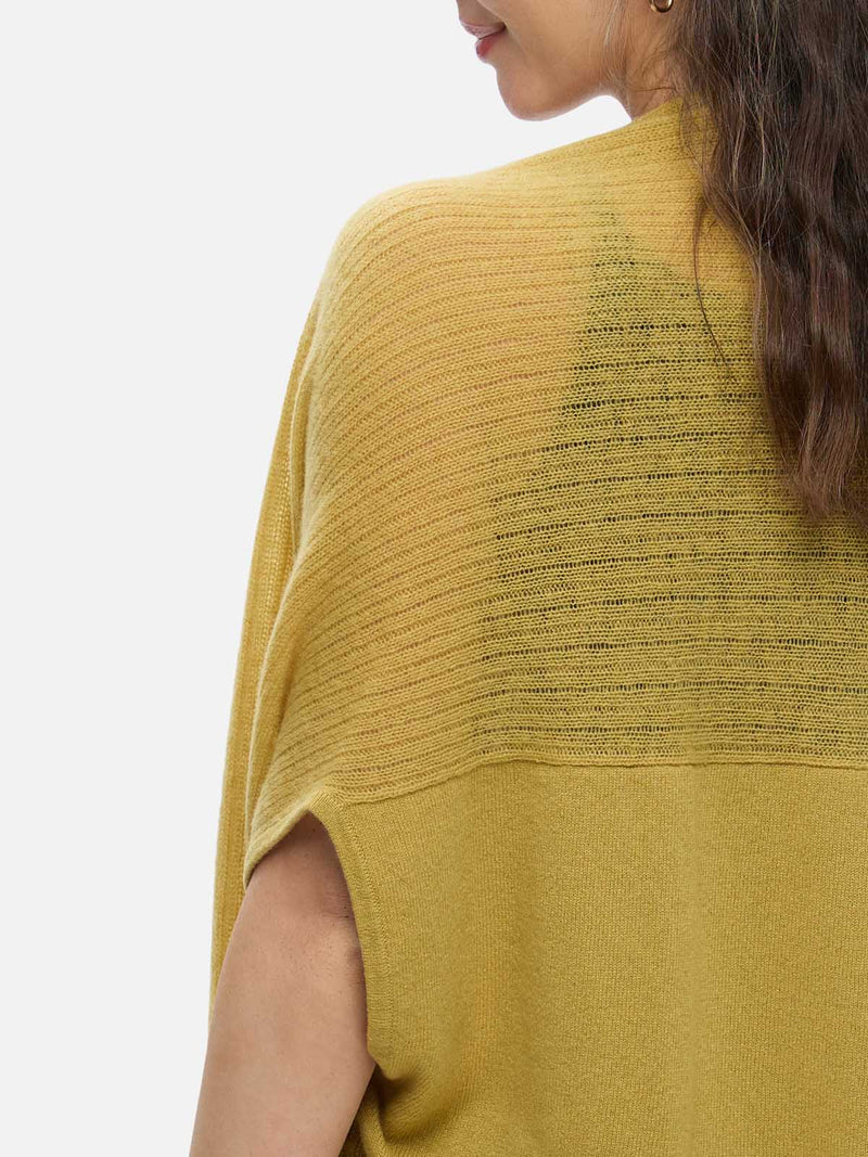 Woman wearing a mustard yellow cashmere vest over a black top and blue jeans on a white background