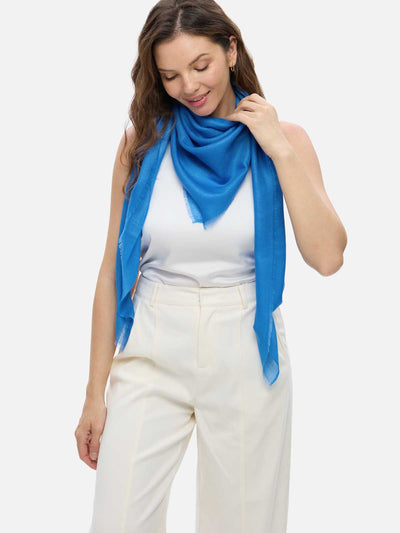 A woman wearing a blue Featherlight Square Cashmere Scarf draped over a white sleeveless top and cream-colored trousers, photographed against a light gray studio background.