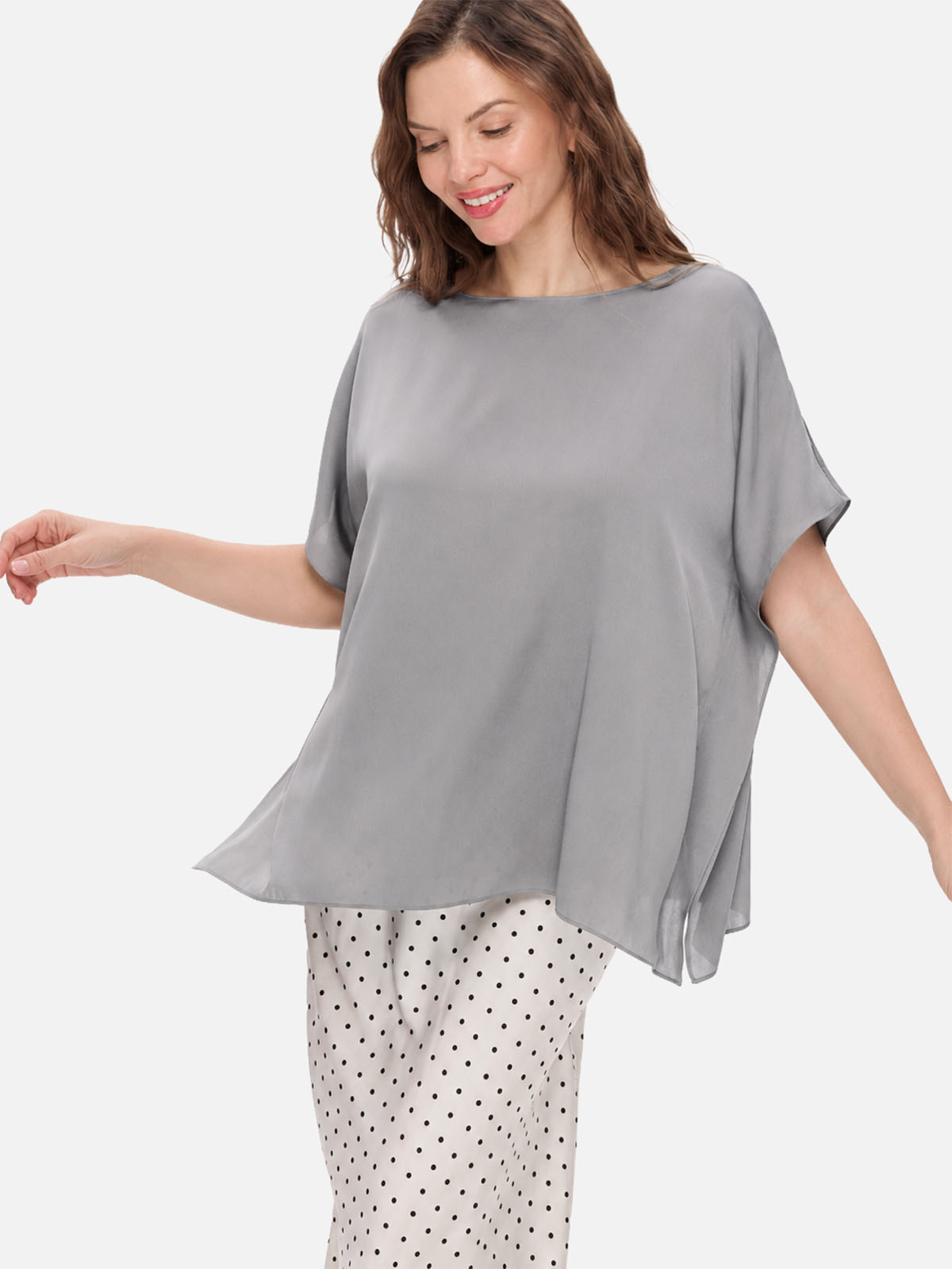 A woman wearing a loose, short-sleeve gray silk top and white pants with black polka dots, photographed against a plain light gray studio background.