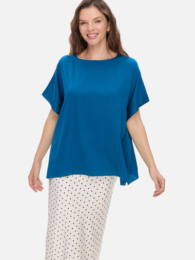 A woman modeling a short-sleeve boat-neck silk top in a vibrant teal blue, paired with white pants featuring a black polka-dot pattern, against a solid light gray background.
