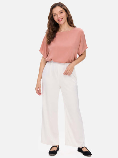 A woman modeling a dusty rose short-sleeve silk top and white wide-leg pants against a light gray studio background.