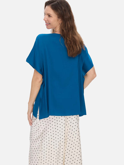 A woman shown from the back wearing a loose-fitting teal blue short-sleeve silk top and white pants with black polka dots, against a light gray studio background.