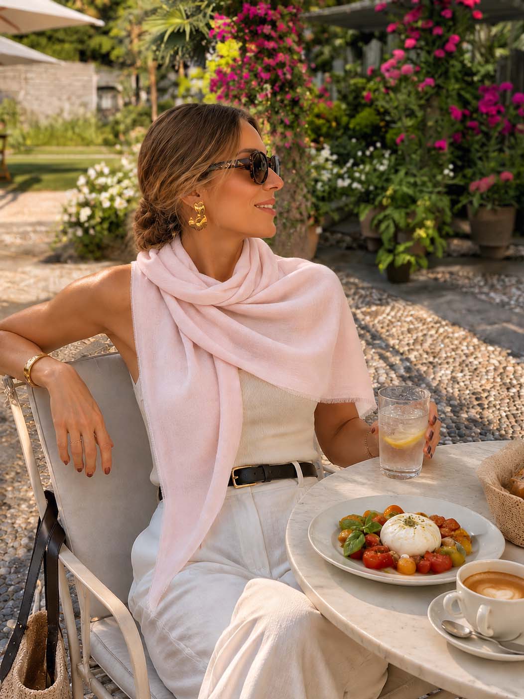 A woman sits at an outdoor patio table wearing a pink featherlight cashmere scarf draped over her shoulders, a white sleeveless top, and white pants. A plate of fresh burrata with tomatoes, a glass of water, and a cup of coffee are on the table in front of her.