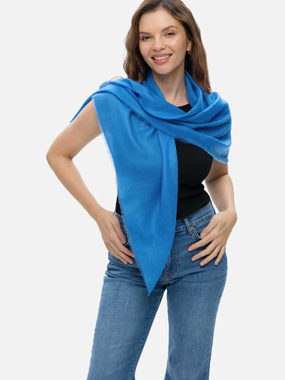 A woman wearing a blue Featherlight Square Cashmere Scarf draped over a black sleeveless top and blue jeans, photographed against a light gray studio background.