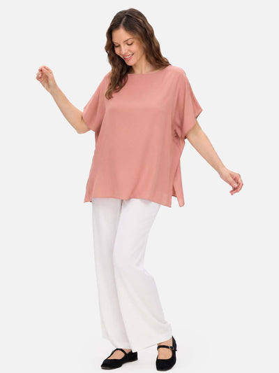 A woman modeling a loose, short-sleeve dusty pink silk top paired with white wide-leg pants and black flats, set against a solid light gray background.