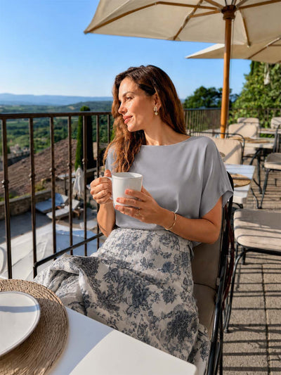 A woman sits on an outdoor patio chair holding a white mug, wearing a light gray silk top and a gray-and-white floral skirt. She is seated under a large cream patio umbrella with a scenic landscape and blue sky in the background.