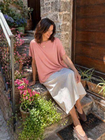 A woman sits on rustic stone steps outdoors, wearing a loose dusty pink short-sleeve silk top and a light beige satin midi skirt. The scene is decorated with potted pink flowers and green plants next to a wooden door.