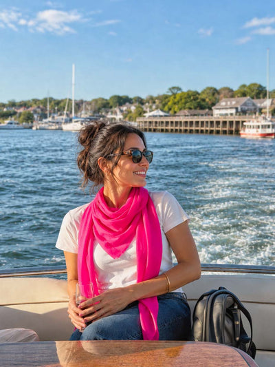 Woman wearing a fuchsia Square Featherlight Cashmere Scarf with a white T-shirt and jeans, relaxing in sunglasses aboard a boat with a sunny harbor in the background.