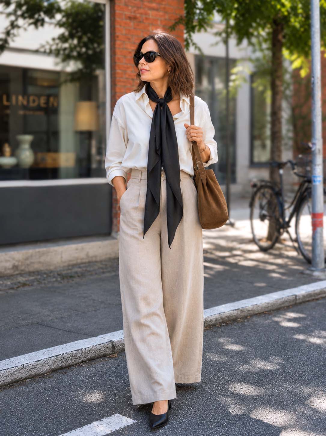 A woman stands on a city sidewalk wearing a cream button-up shirt, wide-leg beige trousers, a black cashmere scarf, black sunglasses, and black pointed flats, while carrying a brown shoulder bag.