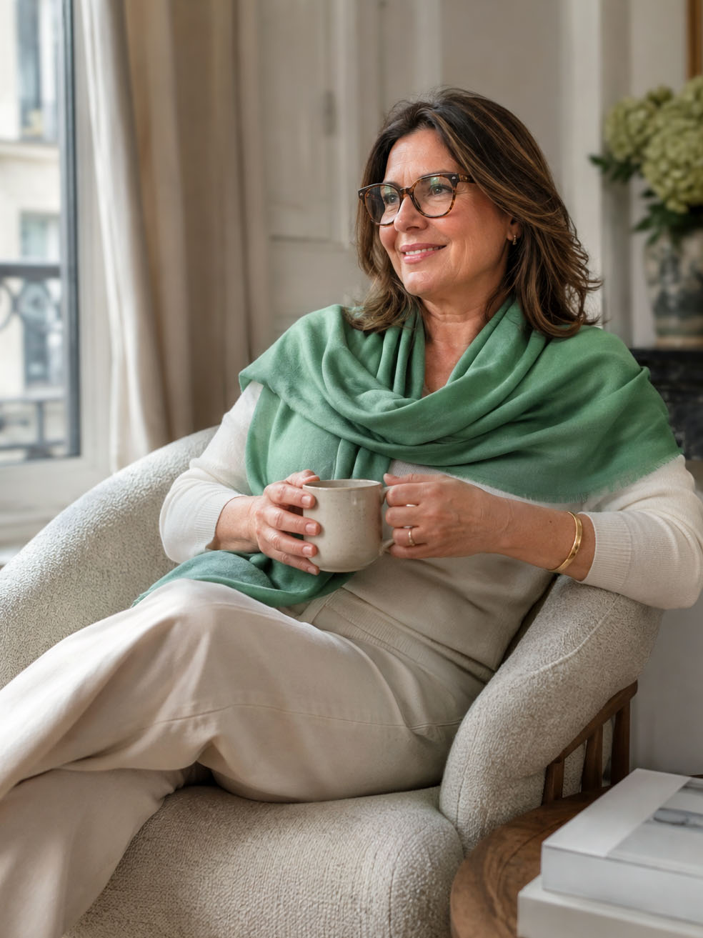 A woman wearing a sage green cashmere scarf draped over her shoulders, sitting on the sofa, holding a cup of coffee.