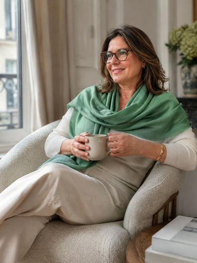 A woman wearing a sage green cashmere scarf draped over her shoulders, sitting on the sofa, holding a cup of coffee.