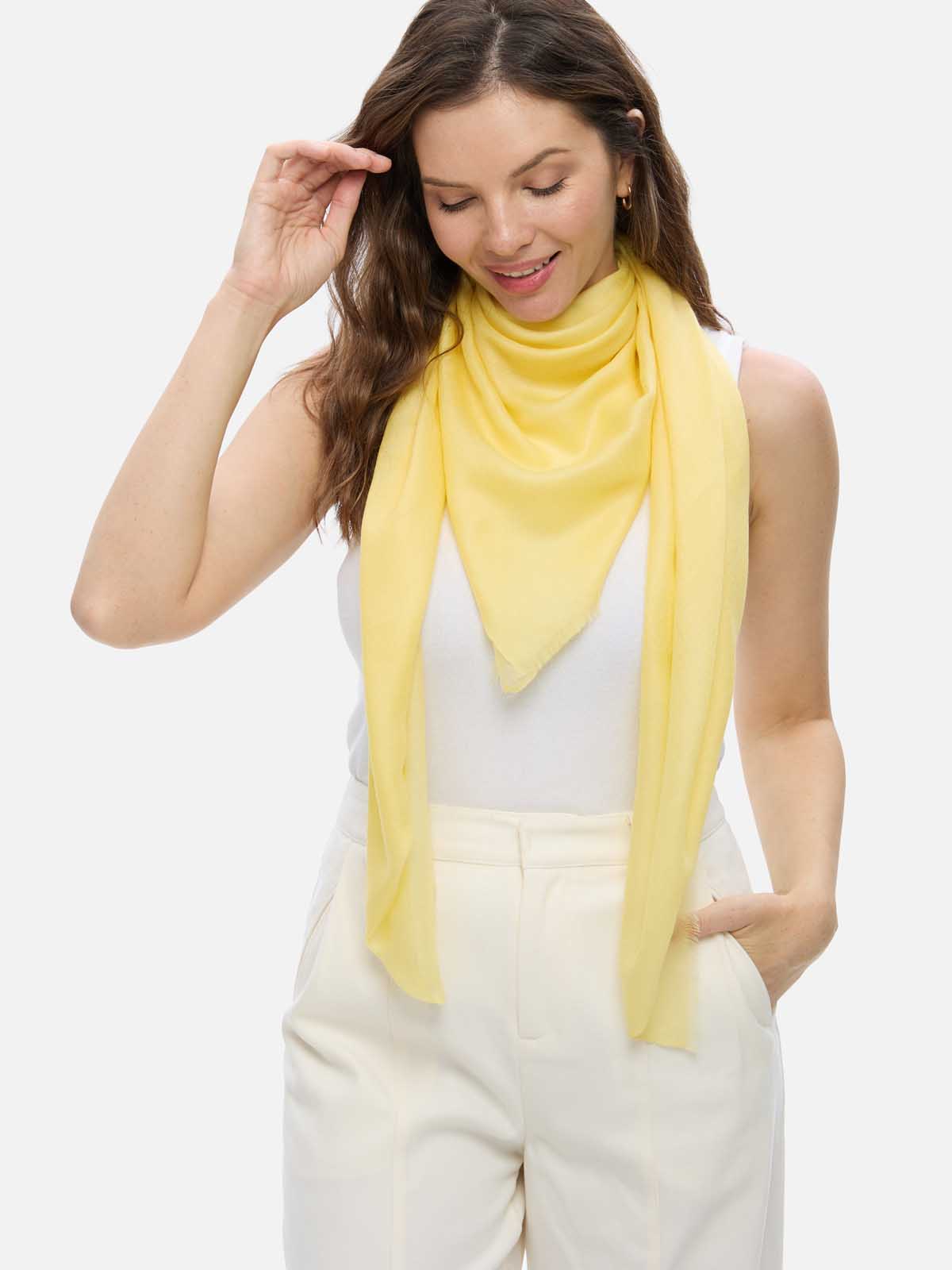 A woman modeling a Featherlight Square Yellow Cashmere Scarf draped over her shoulders, styled over a white top and cream-colored trousers, against a solid light gray background.