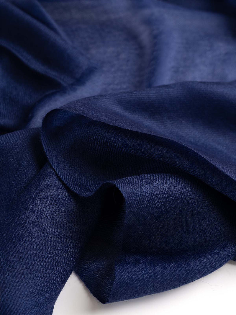 A woman modeling a navy blue Featherlight Square Cashmere Scarf draped over her shoulders, worn over a white top and white trousers, against a plain light gray studio background.