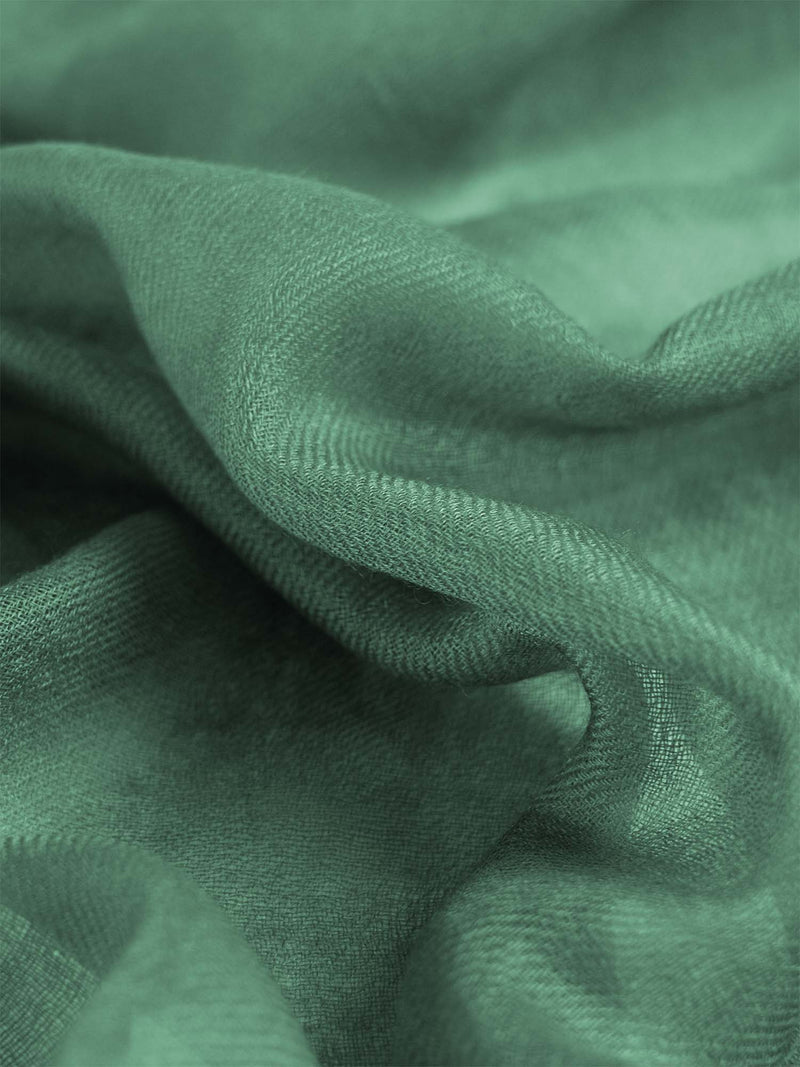 A woman wearing a sage green cashmere scarf, paired with a white top and cream-colored trousers, against a solid light gray background.